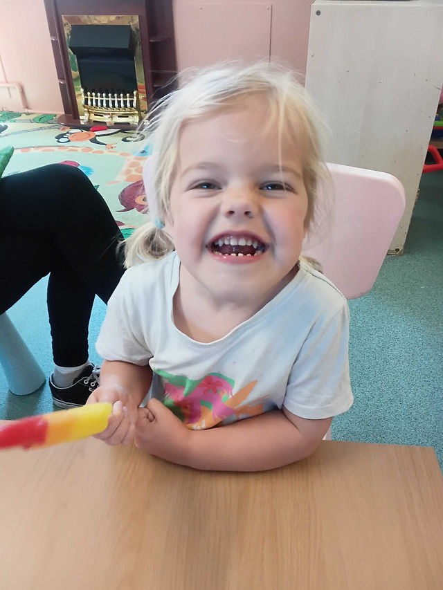 blonde haired little girl is smiling widely in the camera holding an ice lolly in her hand