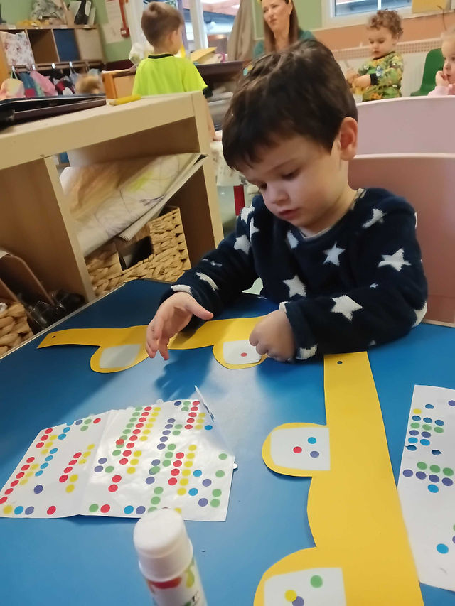 a little boy is sitting by a blue table, there is a yellow card, shaped like Pudsey ears, he is sticking tiny little dot stickers on the ears