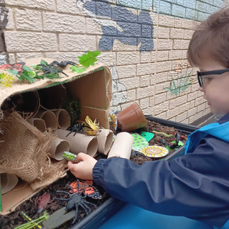 a child wearing glasses puts toy bugs into a bug hotel made of card board, tubes, soil and leaves