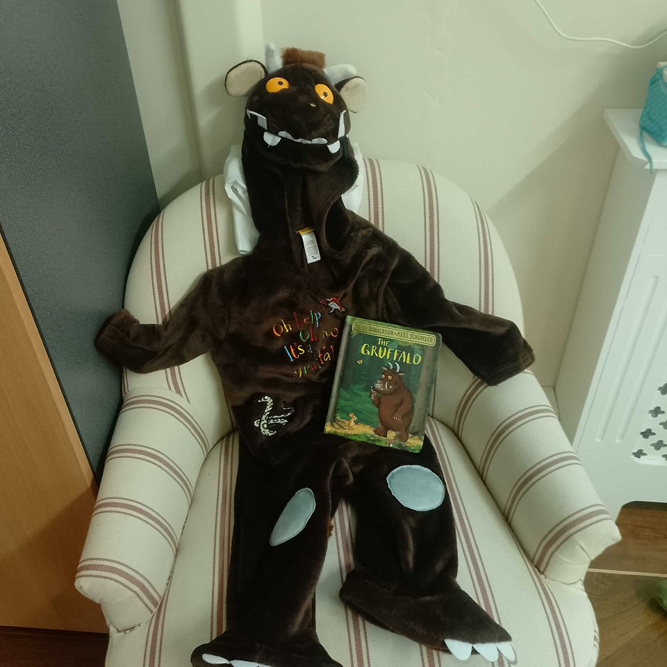 The Gruffalo is sitting on a light coloured armchair with the same titled story book in his lap