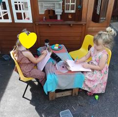 two little girls are stiiting on yellow chairs, making marks on paper that is attached to a clipboard, there is small table with a tablecloth between them, in the background there is a wooden playhouse