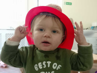 a baby wearing a red plastic firefighter helmet, holding onto it with both hands, looking at the camera