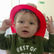 a baby wearing a red plastic firefighter helmet, holding onto it with both hands, looking at the camera