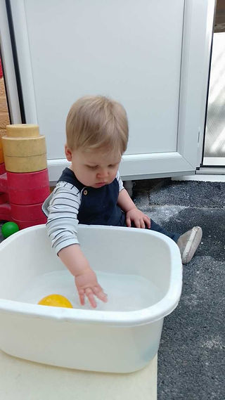 baby boy is sitting next to a white bowl splashing water with his hands