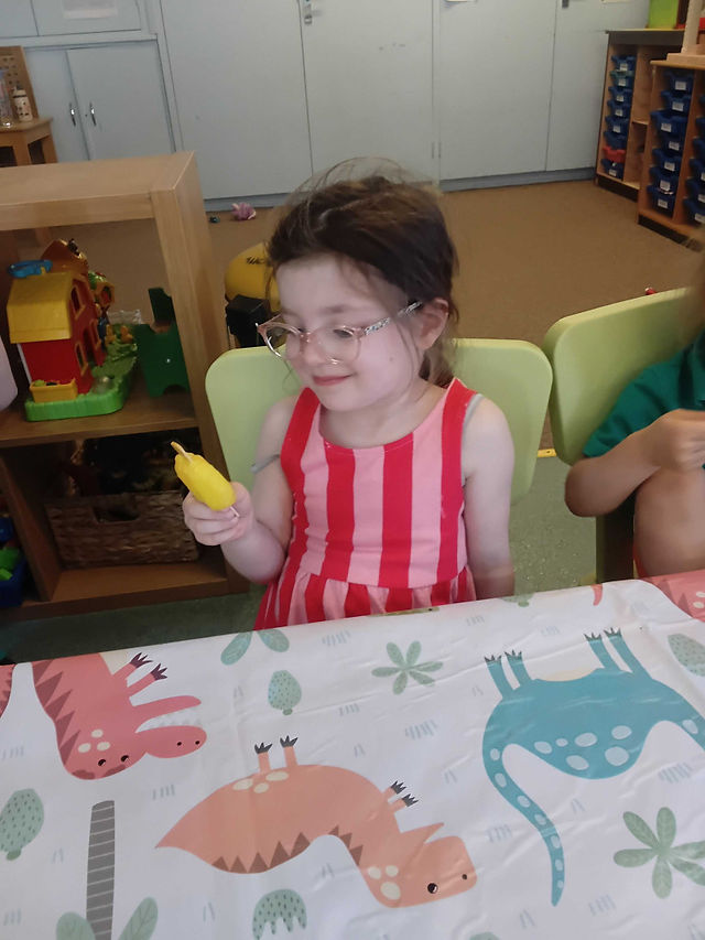 a broun haired little girl, wearing glasses and a stripey dress is looking at the ice lolly in her hands, she is smiling happily, in the background there is a unit with toys and a carpet