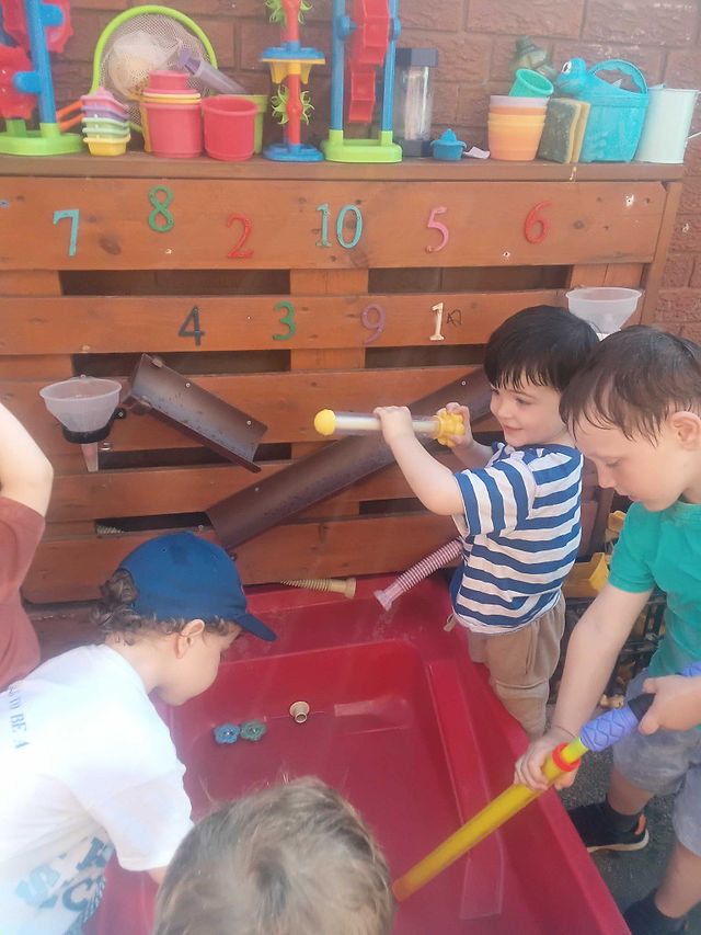 a group of young children play happily with big water syringes, spraying each other with water over a large red water tray and a wooden water wall