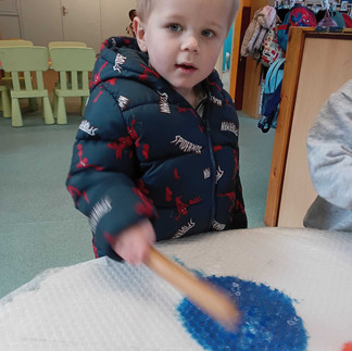little boy is using a rolling pin on bubble wrap to spread blue paint at nursery in Totton