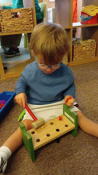 a little toddler wearing glasses is uing a toy hammer to push pegs in a board