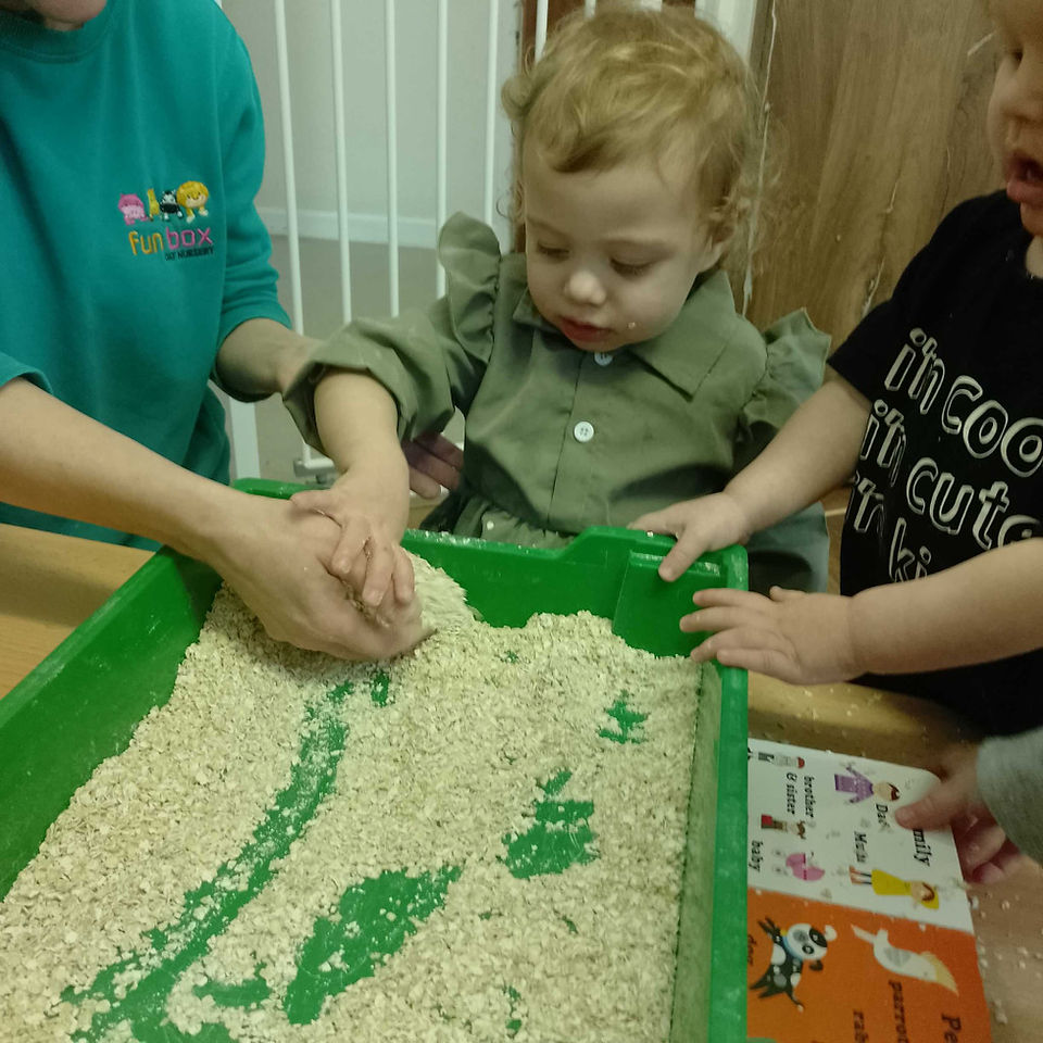 two young children exploring a green tray full of oats with the help of a teacher who is wearing Fun Box Day Nursery uniform