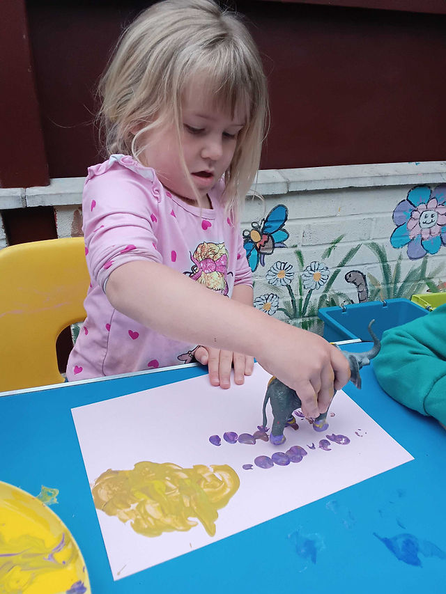 little girl in a pink top is holidng a toy animal dipped in paint to make marks on paper