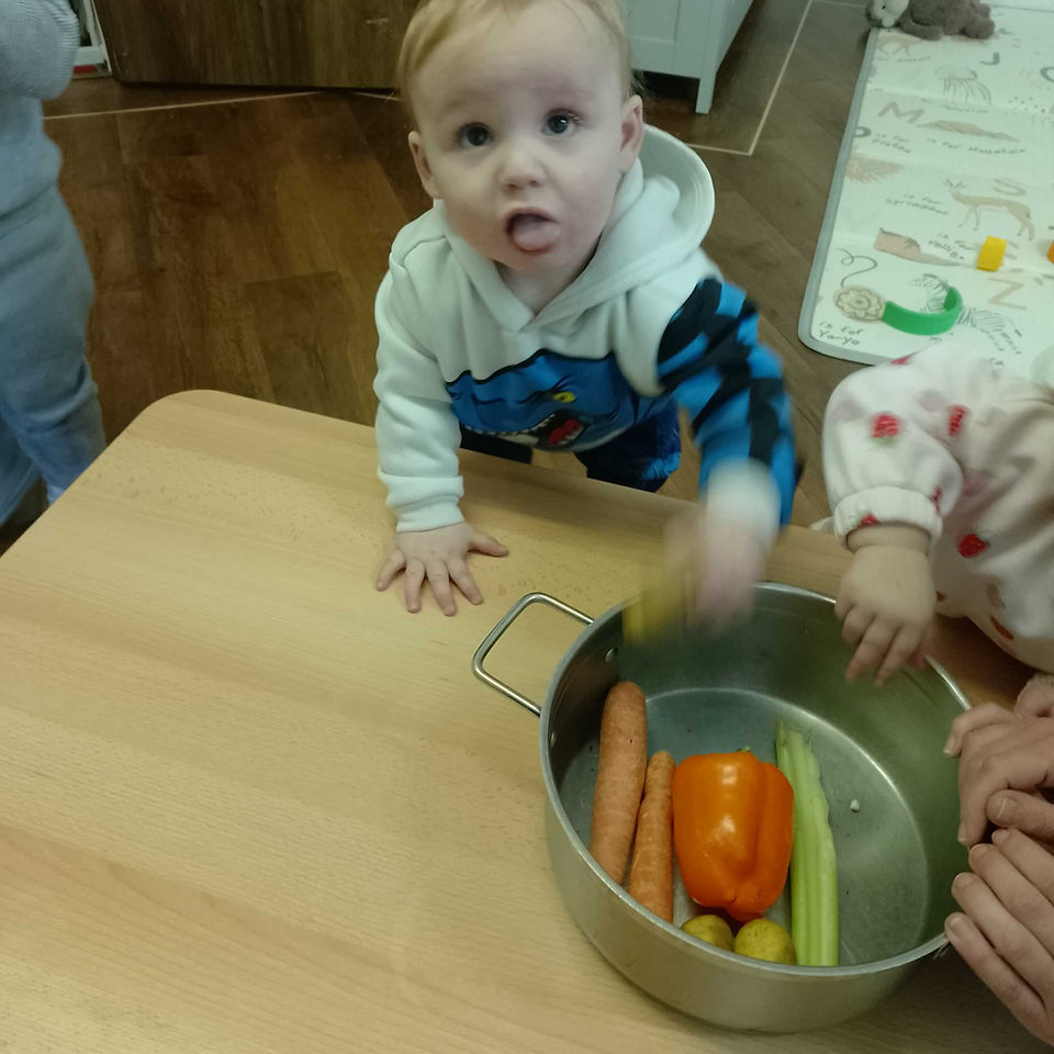 a baby boy looking into the camera, big pot of various vegetables are in front of him on a low table