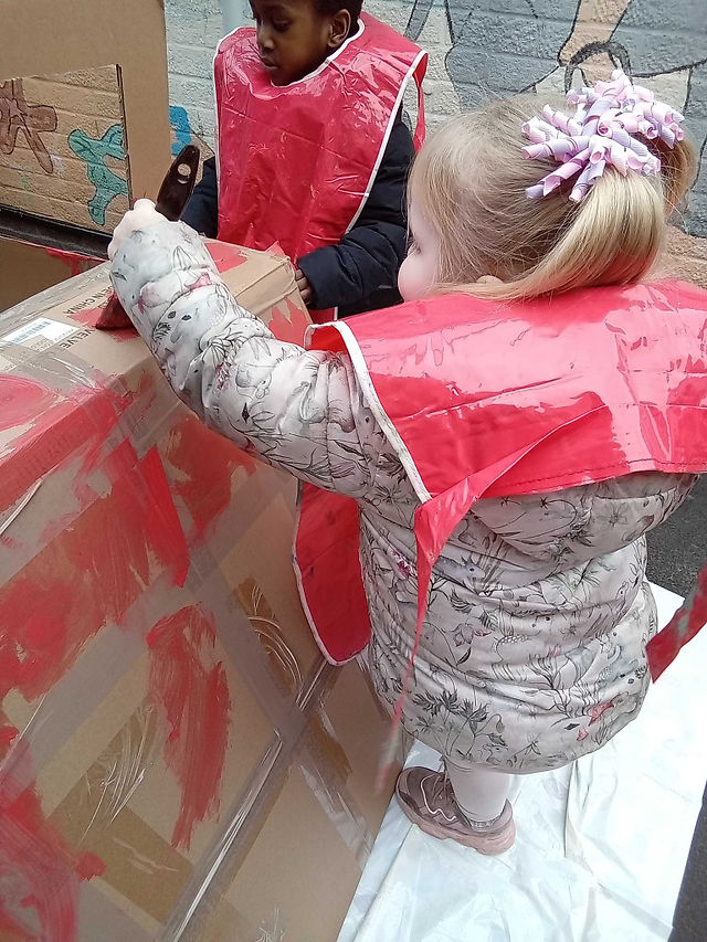 Little girl wearing a pink jacket and a red tabard is painting a large cardboard box to red