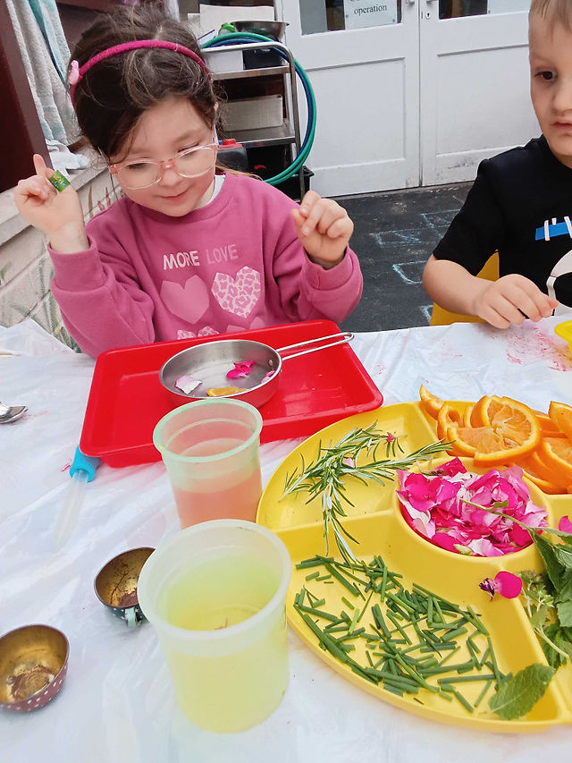a little girl wearing pink top and glasses is mixing herbs and fruits in a little metal pot during a nursery activity
