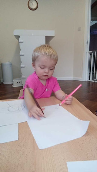 a baby boy wearing a pink t-shirt is holding a pencil in each hand and drawing on a piece of paper