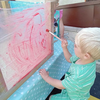 a blonde haired child is painting with red paint on a vertical, see through surface, another child is in the background