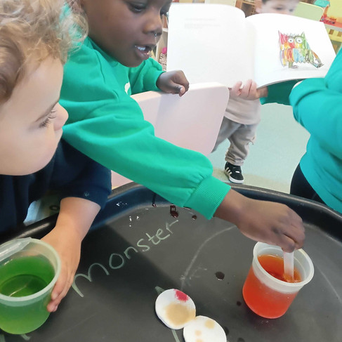 two children a drawing coloured water from a tub using a pipetter to put on the white cotton pads in a sensory tray in front of them
