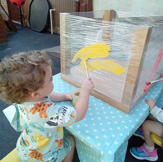a curly haired child is using a paintbrush to paint on cling film streteched across two wooden poles, nursery playground behind him