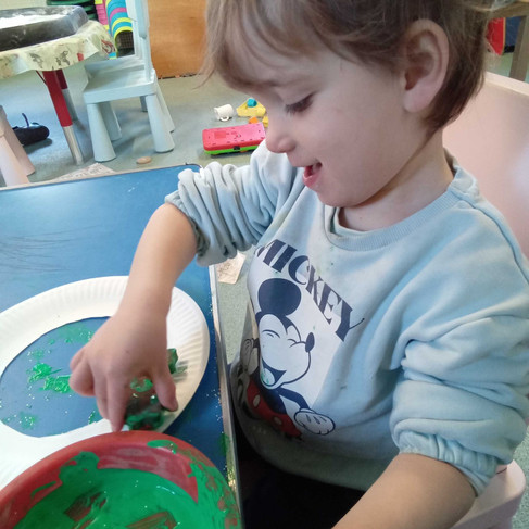 a little boy is smiling as he is printing with green paint and Christmas themed stamps onto a cut out paper plate