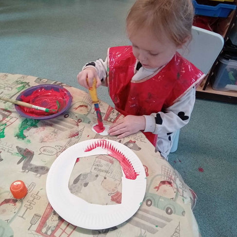 a little girl, wearing a red apron is painting a cut out papaer plate with red paint