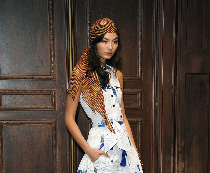 Slender model wearing a brown yellow and orange printed side knot head scarf standing in front of a dark varnished wood door.