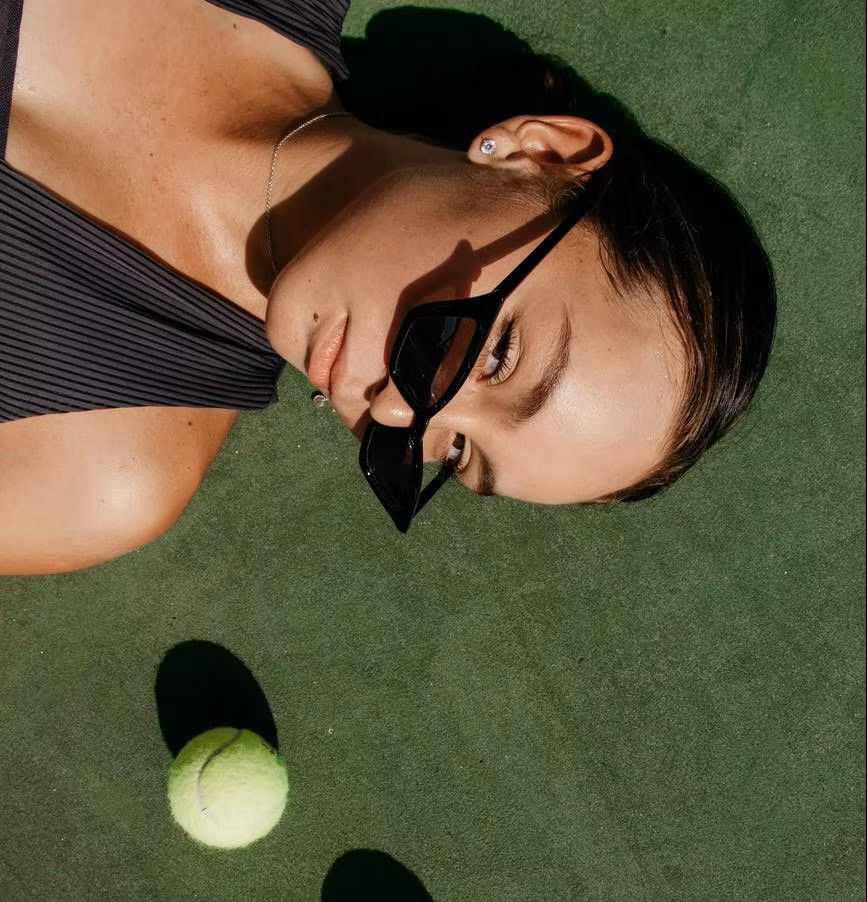 Woman lying on a tennis court wearing sunglasses with two tennis balls next to her head.