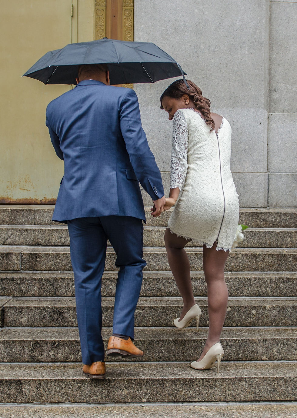 A black couple on their wedding day outside of city hall. Husband in a navy blue suit and brown suit. Wife in a DVF long sleeved, zipper back, off white dress. He holds an umbrella to protect them from the rain.