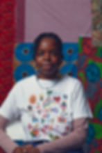 A young Black woman with canerowed hair, wearing a white t-shirt with colourful emblems and the words 'Trying my Best' across the top, and underneath a lilac long sleeved t-shirt. She wears long earrings and two necklaces. She looks straight at you, with a slight smile. The background is a collage of African batik fabrics