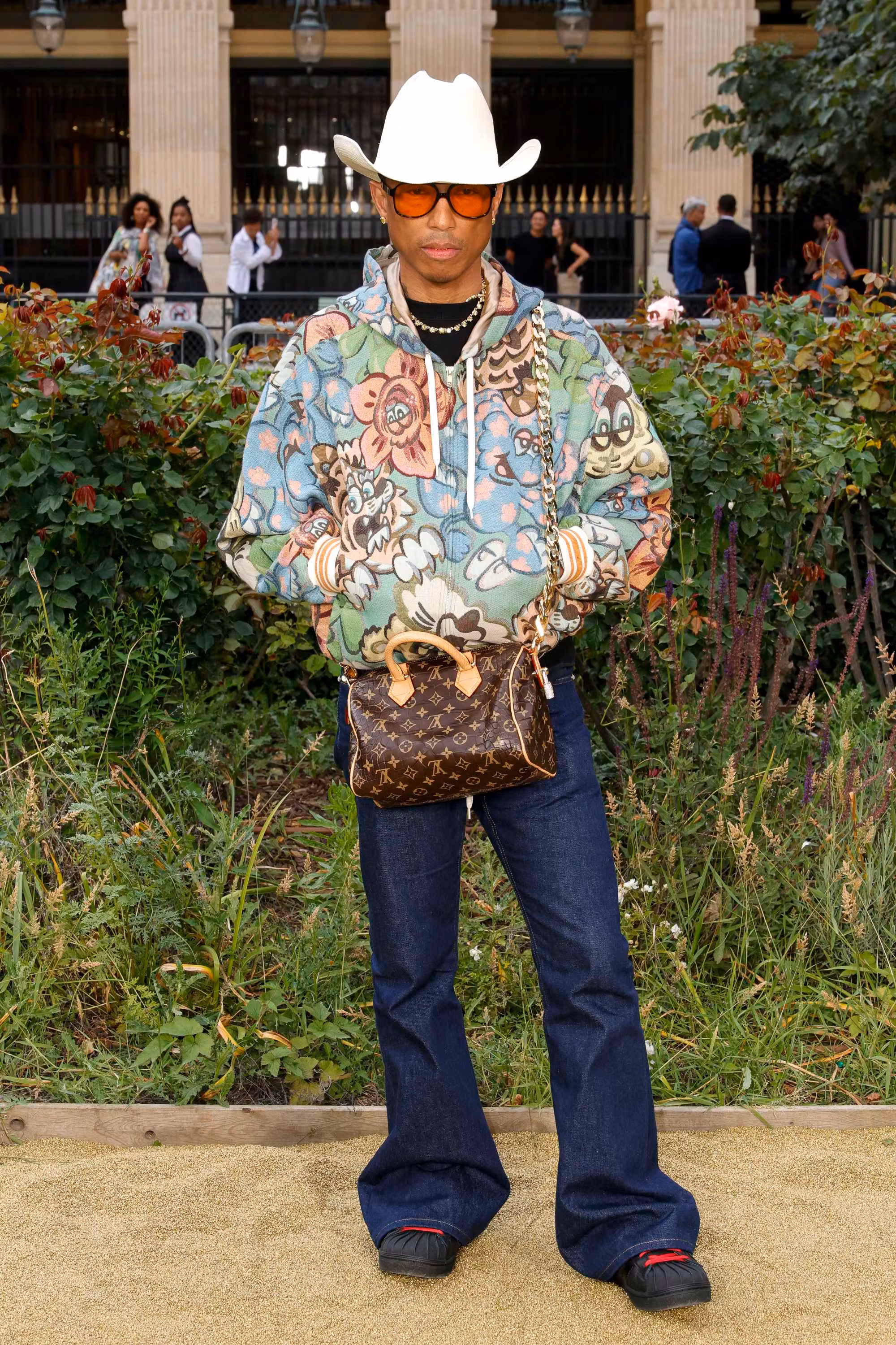 Pharrell Williams poses in floral tapestry hoodie, white cowboy hat, amber sunglasses, dark denim flares and a Louis Vuitton Speedy bag, greenery behind.