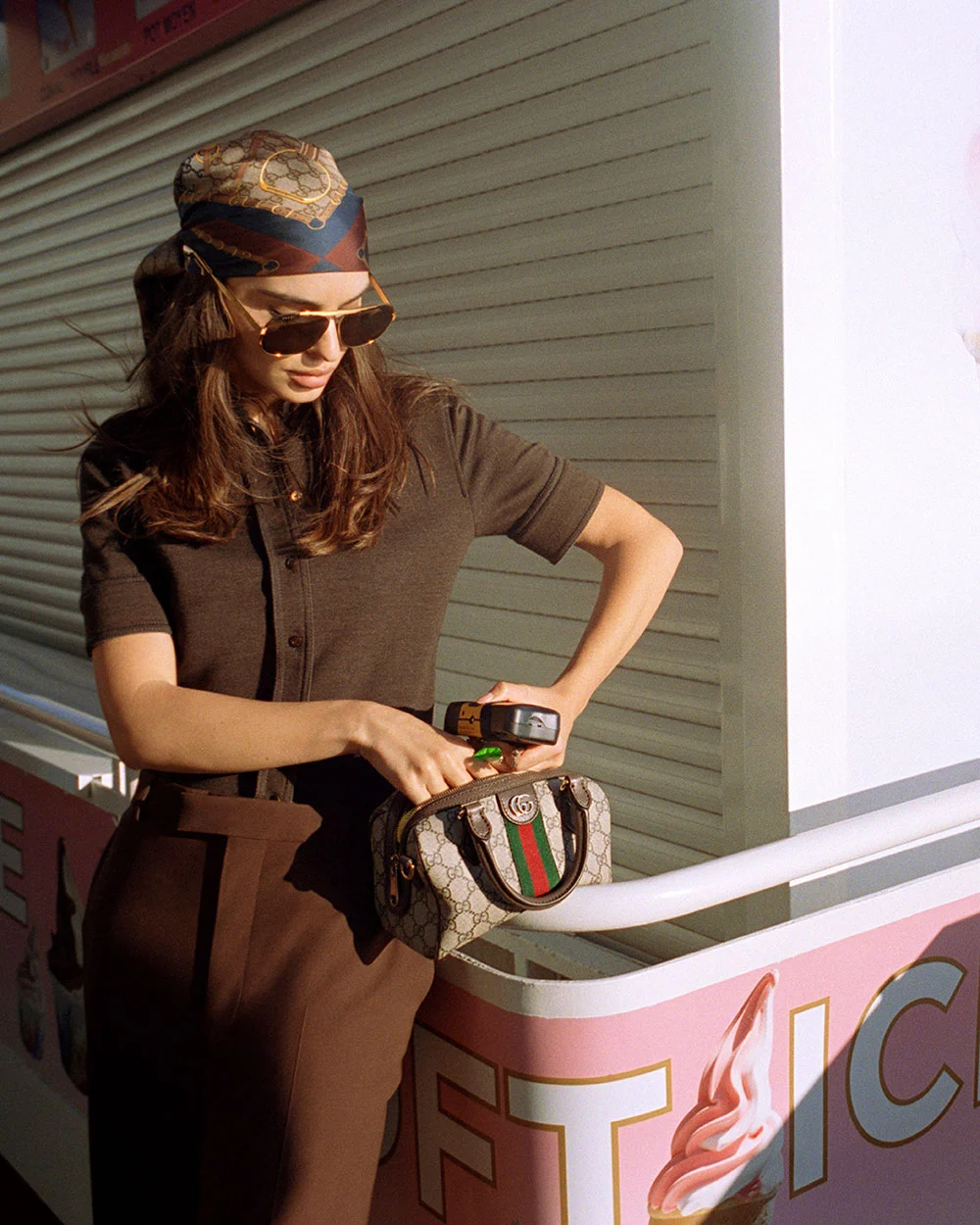 Emily Ratajkowski wearing a headscarf, sunglasses, and short-sleeved knit top, standing near a pastel ice cream stand while opening the Gucci Giglio handbag with green and red web stripe. Shot in soft daylight.
