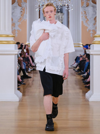 A model walks a runway in a white short-sleeved shirt with ruffled edges, black shorts, and black platform Crocs, clutching a large, white pillow-like bag with a ruffled trim
