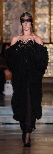A model walks a runway wearing a large, heavily textured black off-the-shoulder wrap piece, paired with a black skirt and a sheer black visor