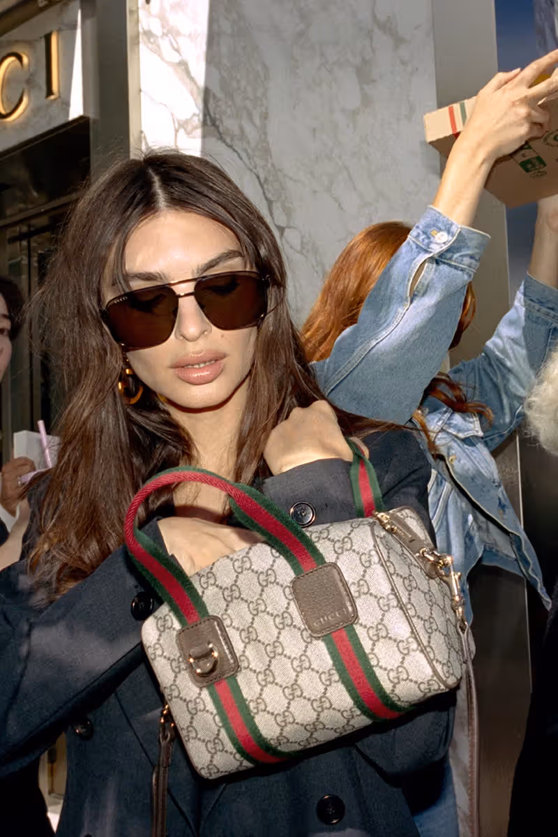 Close-up of Emily Ratajkowski wearing dark sunglasses and hoop earrings outside a Gucci storefront, holding the Giglio handbag with pink webbing, surrounded by a crowd in motion. Flash-lit, candid style.