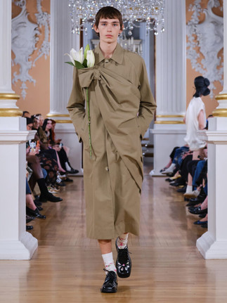 A model walks a runway in a khaki trench coat that is asymmetrically draped and knotted across the chest, with a large white lily tucked into the knot