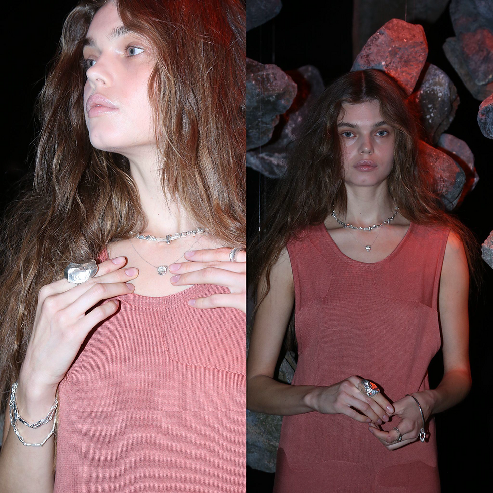 A model with bleached blonde hair kneels by a pool of water in a darkened, rocky setting, holding a large, reflective silver sphere and a delicate chain