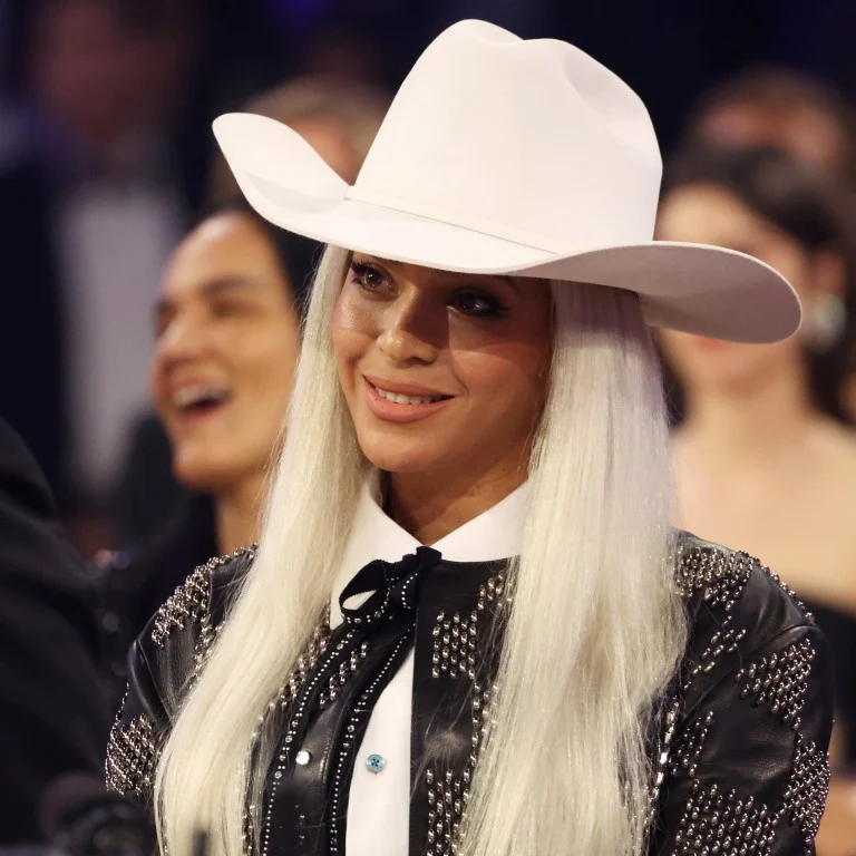 Beyoncé smiles in studded black leather jacket, long platinum hair and crisp white cowboy hat, blurred audience reacting in background.