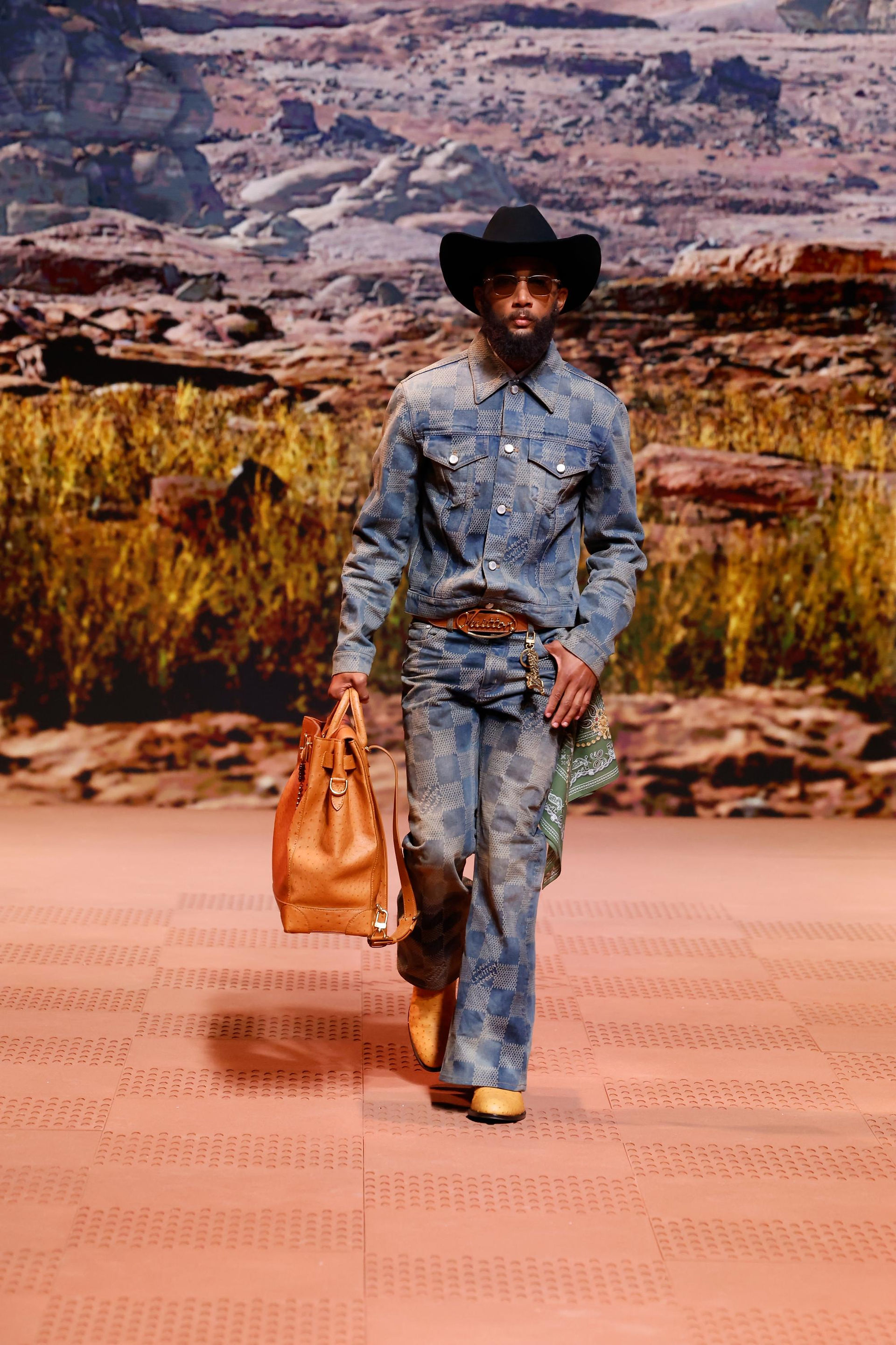 Model walks on terracotta runway wearing matching checkerboard denim jacket and flared jeans, wide black cowboy hat, tan ostrich tote and brown belt.