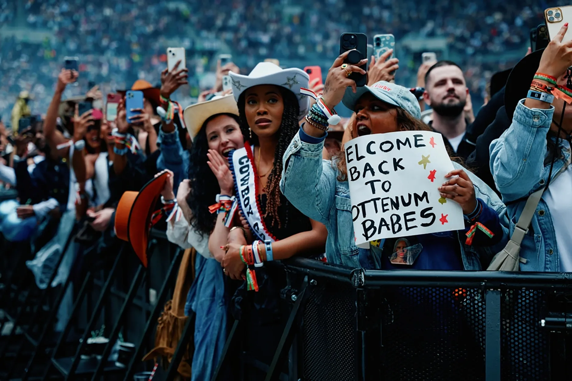 Front-row crowd shot of women in cowboy hats and denim jackets cheering, one holding a handmade sign reading “Welcome back to Tottenham babes”, phones raised in the air.