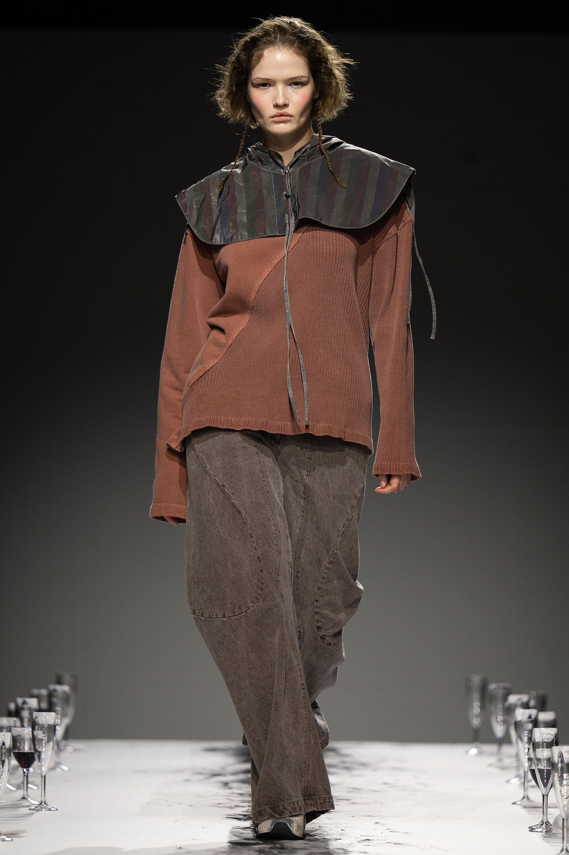 A model walks a runway in a reddish-brown knit sweater with a large, structured collar made of a different material, paired with washed-out baggy trousers