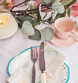 A close up picture of a pink cup and saucer, a china cake plate with butter knife and cake fork, displayed with eucalyptus 