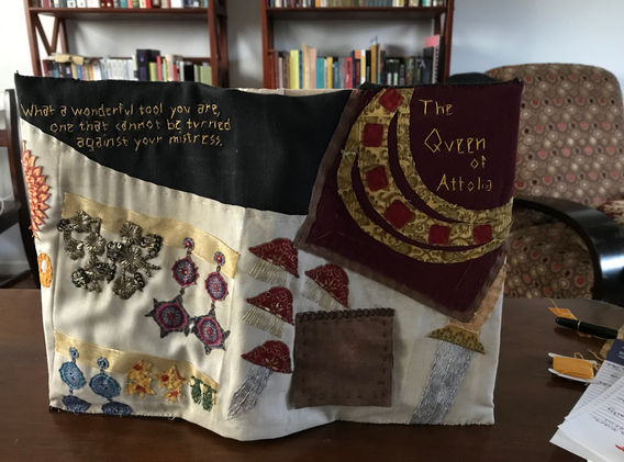Open spread of the full book cover of Queen of Attolia, embroidered to show the queen's traveling jewelry collection of her bespoke royal headpiece, iconic hair clip/dagger combo, bee earrings, and several sets of mismatched earrings given to her by Nahuseresh. On the front cover is an open-able flag for the small box of ruby earrings given by a certain cheeky thief.