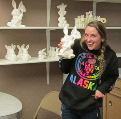 Woman holding bunny, surrounded by ceramic bunnies in a Ceramic Cafe. Alaska hoodie.