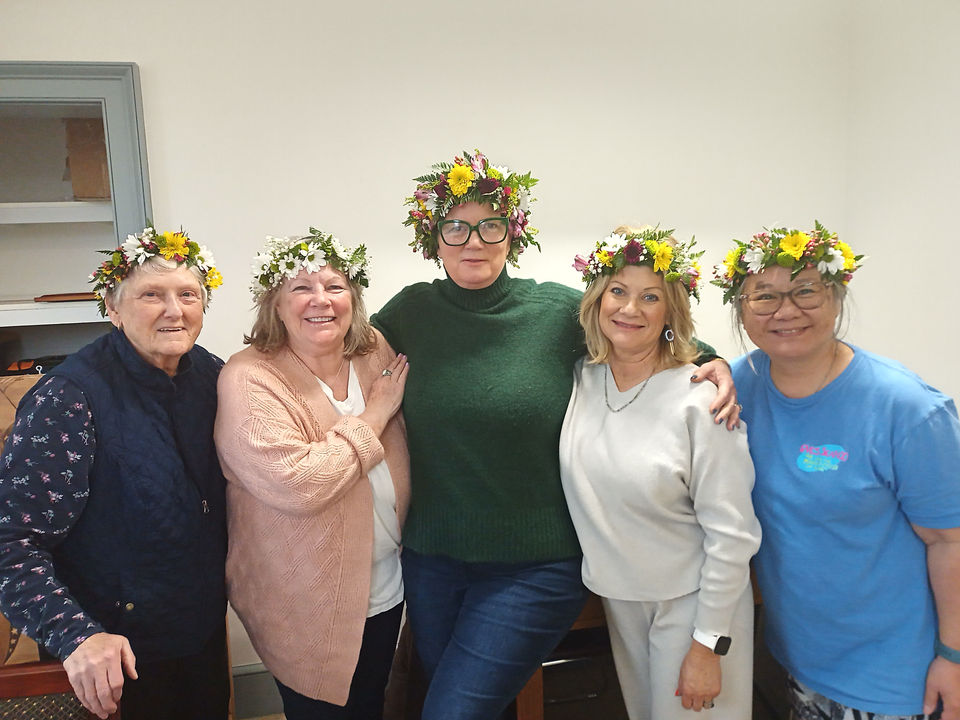 Five women wearing floral crowns, smiling for a photo