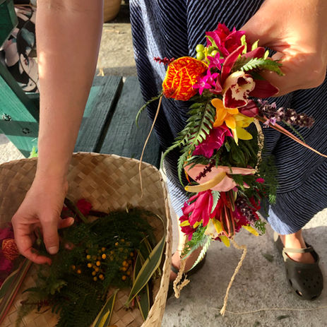 Person arranging colorful flowers and greenery in a basket, with a floral lei in their other hand