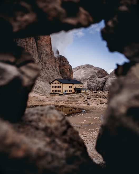 A small mountain hut, nestled among rugged cliffs, framed artistically through an opening in rocks. The hut stands near a shallow puddle, with barren rocky terrain surrounding it, giving a sense of isolation in a dramatic alpine environment