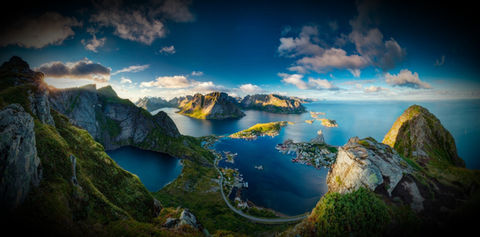 Scenic view of Lofoten, Norway, showcasing dramatic, jagged peaks rising sharply from the deep blue waters of the Norwegian Sea. Traditional red and white Norwegian fishing cabins dot the shoreline, contrasting with the lush greenery of the rugged terrain. The scene captures the unique beauty and remote tranquility of this Arctic Circle archipelago under a clear, expansive sky.