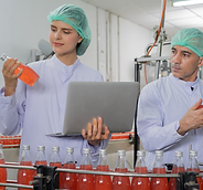 dois funcionários do setor de qualidade, uma mulher e um homem, ambos de jaleco e touca, averiguando a linha de produção de refrigerantes