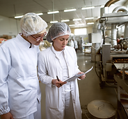 Duas mulheres de jaleco e touca lendo um papel, ao fundo há uma linha de produção de alimentos