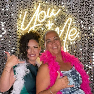Smiling wedding guests in front of silver background with giant "You & Me" neon sign behind them.