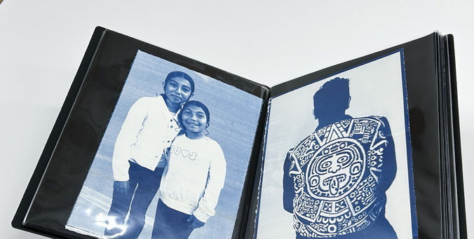 Two arms hold open a black artist portfolio showing two cyanotypes featuring portraits of people.