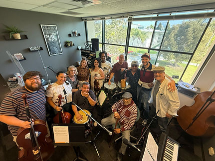 Large group of people wit instruments in a room looking up atthe camera.